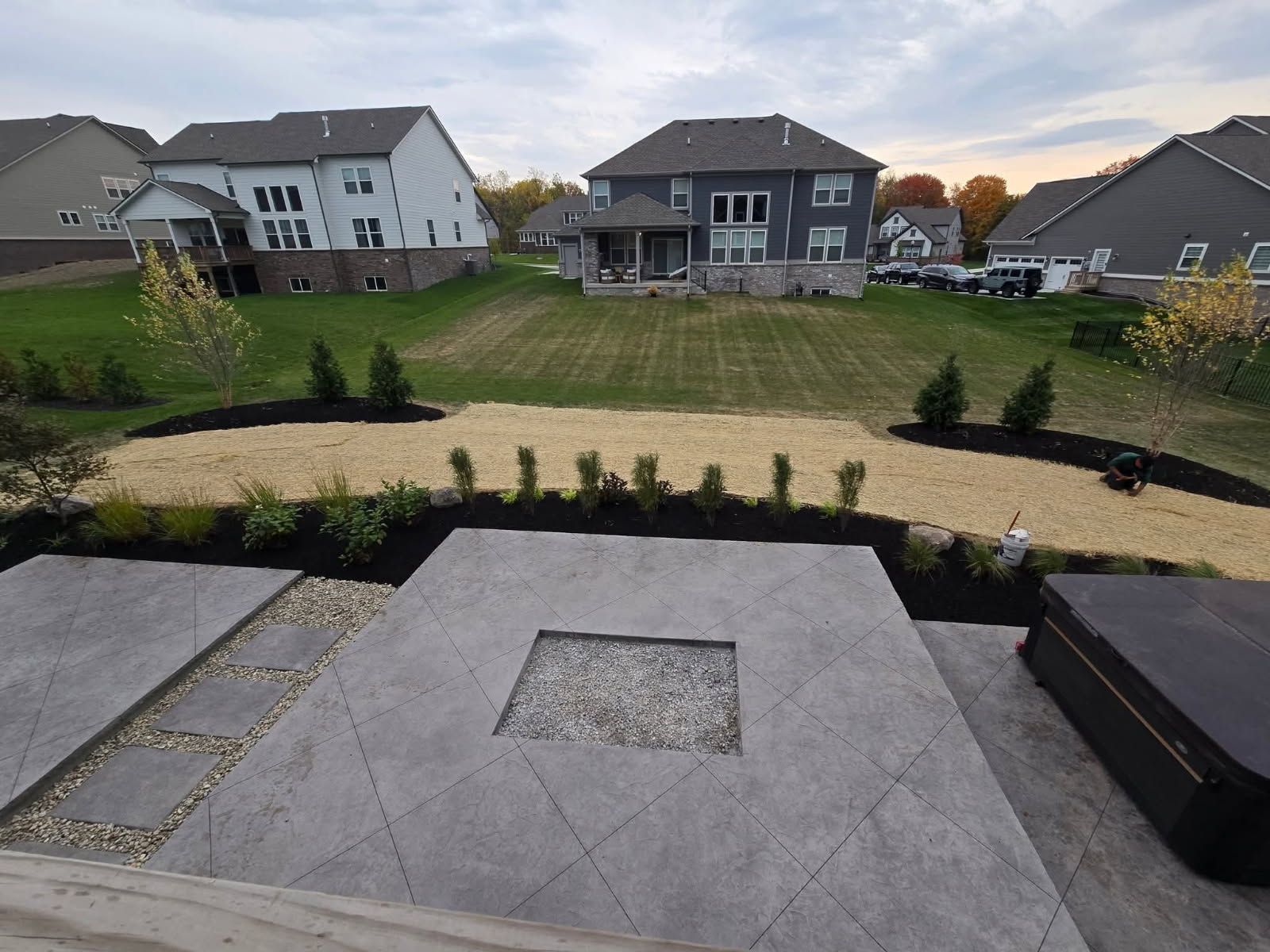 Patio with fire pit, gravel landscaping, and view of houses on a hill with grass, under an overcast sky.