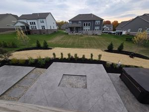 Backyard patio with fire pit, gravel, and grass, with houses in the background under a cloudy sky.