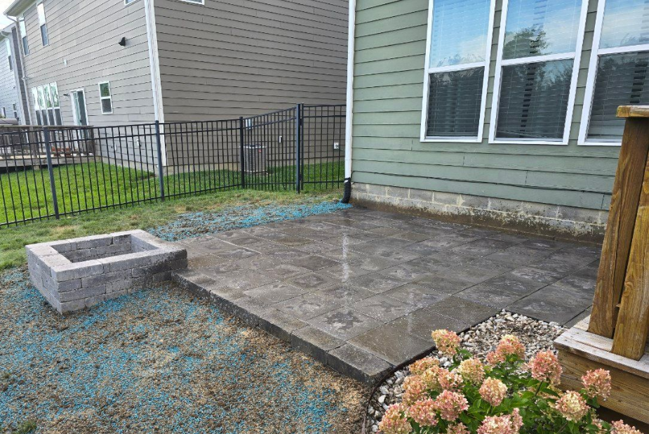 Patio with gray pavers, fire pit, and fence. Green siding on house, grass and flowers present.
