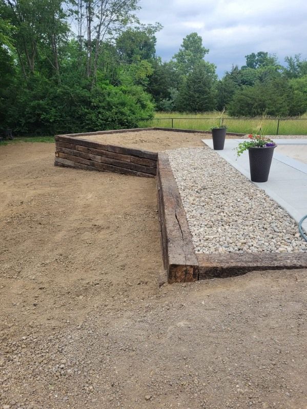 A gravel and soil landscape bed with wooden borders and a dark pot of flowers on a concrete surface.