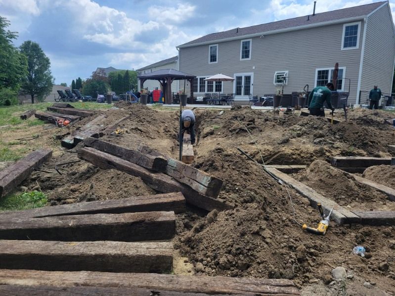 Workers digging in a backyard, using wood and tools for construction near a house with a cloudy sky.