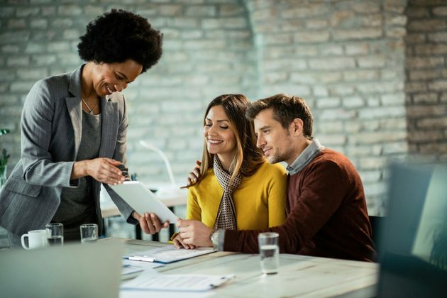 Person in a blazer writing on paper with a silver pen, desk, window in background.