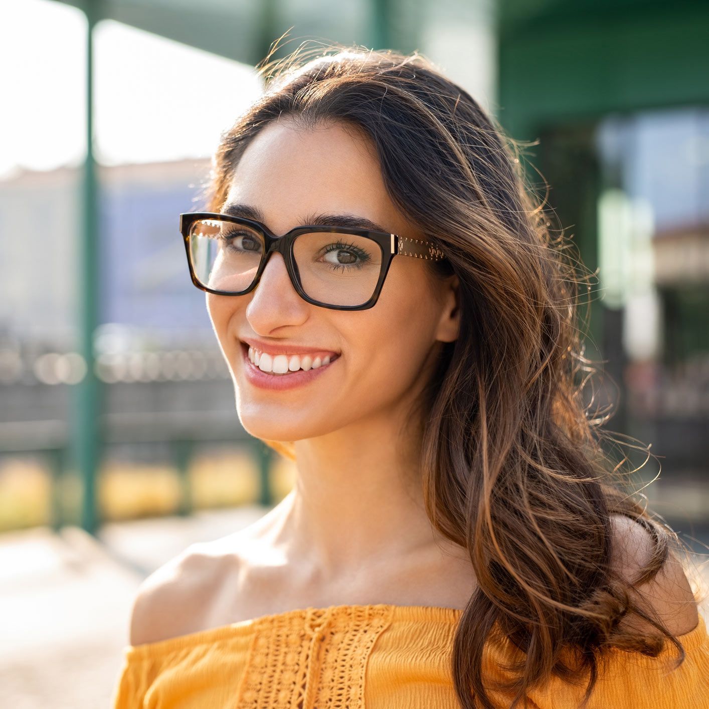 A woman wearing glasses and a yellow off the shoulder top is smiling