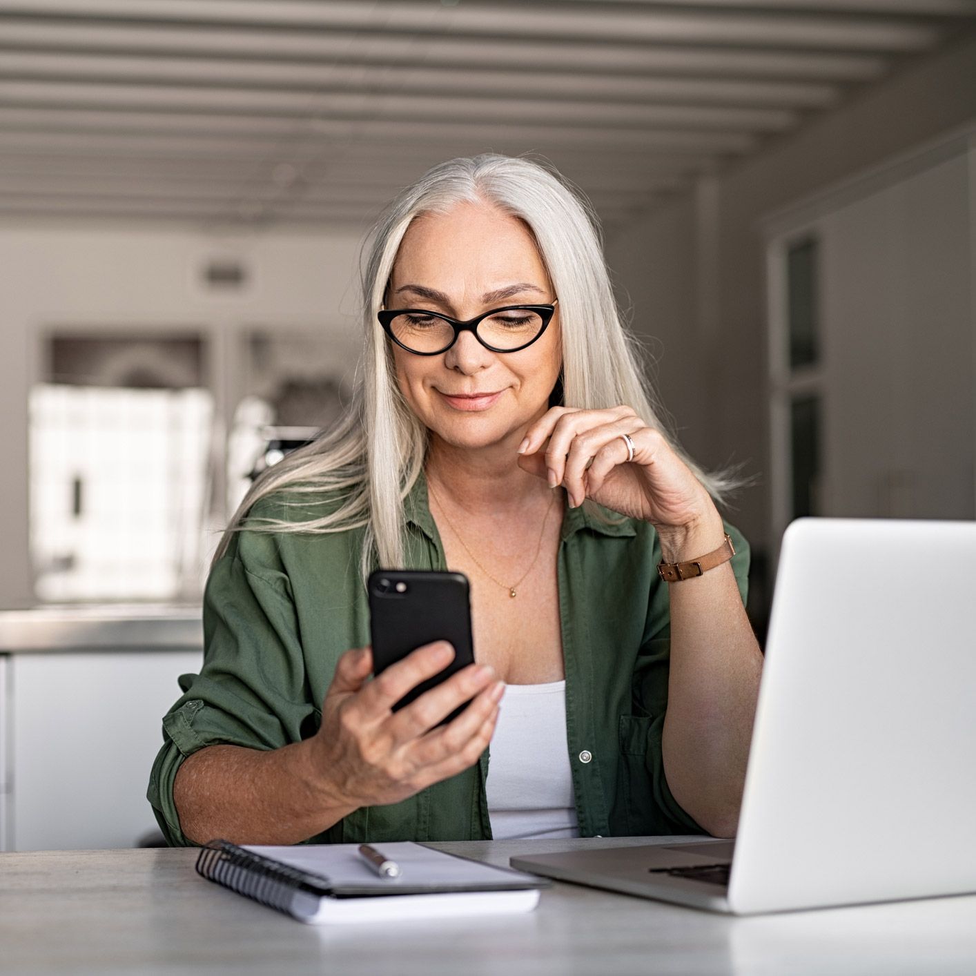 A woman is sitting at a table using a laptop and a cell phone