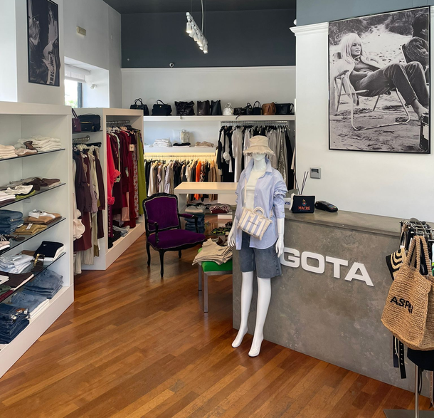 Piccola boutique di abbigliamento con scaffali pieni di vestiti colorati, un manichino e un bancone con la scritta