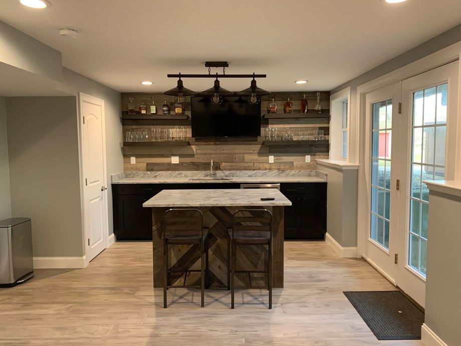 A modern home bar with dark cabinets, a stone island with two stools, rustic wood shelving, and French doors.