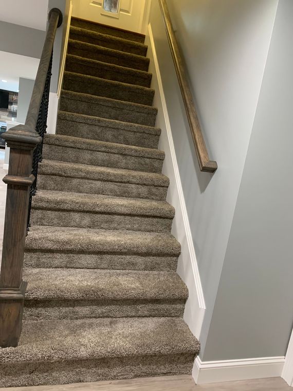 A straight staircase with brown carpeted steps, a dark wood banister on the left, and a wooden handrail on the right wall.