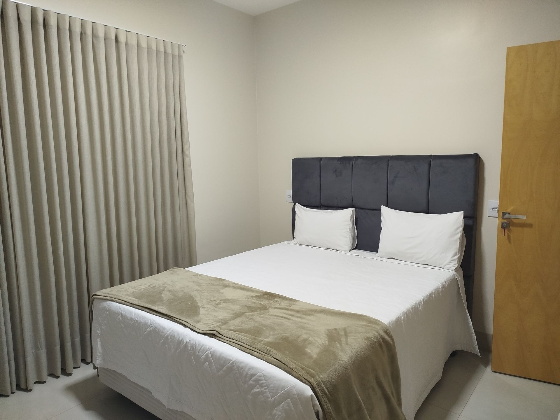 Quarto com cama de casal, cabeceira cinza, roupa de cama branca, manta bege e porta de madeira.