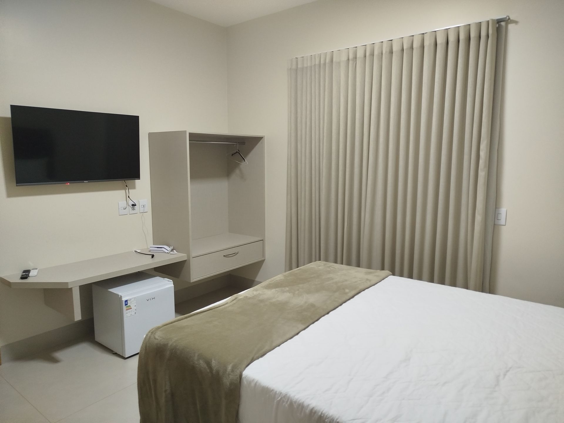 Um quarto de hotel com cama, TV, armário, escrivaninha e frigobar. Paredes claras, cortinas e colcha bege.