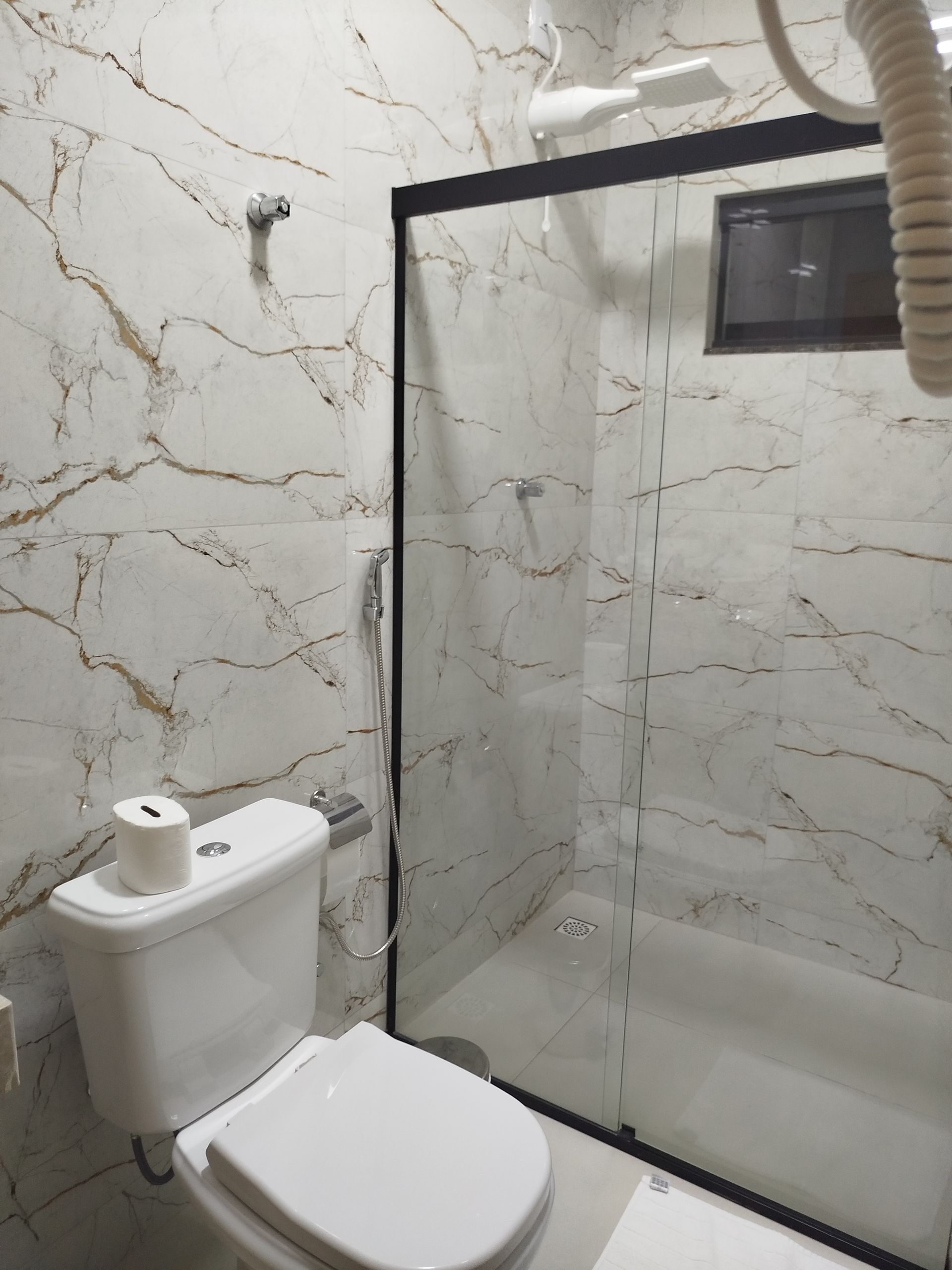 Banheiro com vaso sanitário branco, chuveiro de vidro e paredes com padrão de mármore.