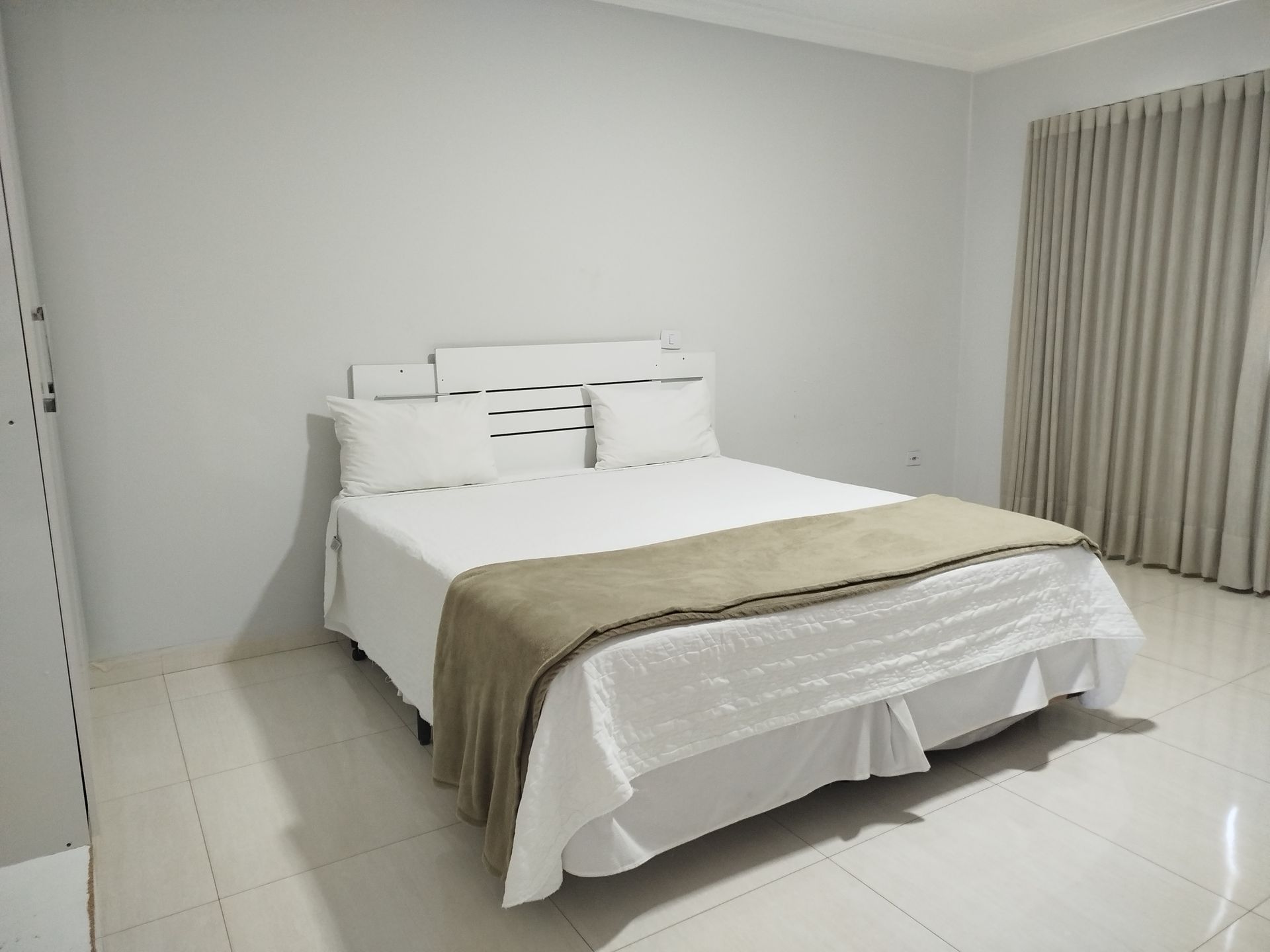 Quarto com cama branca, manta bege, travesseiros brancos, paredes e cortinas cinza.