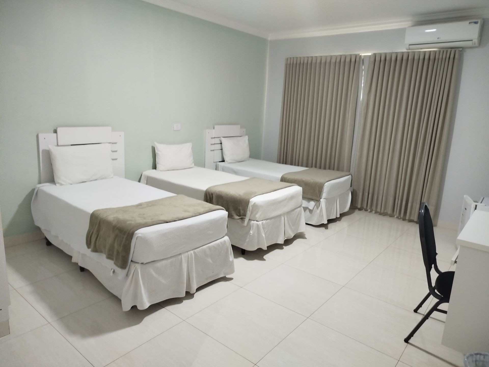 Três camas de solteiro em um quarto com paredes e cortinas neutras; uma escrivaninha e uma cadeira ficam no canto.
