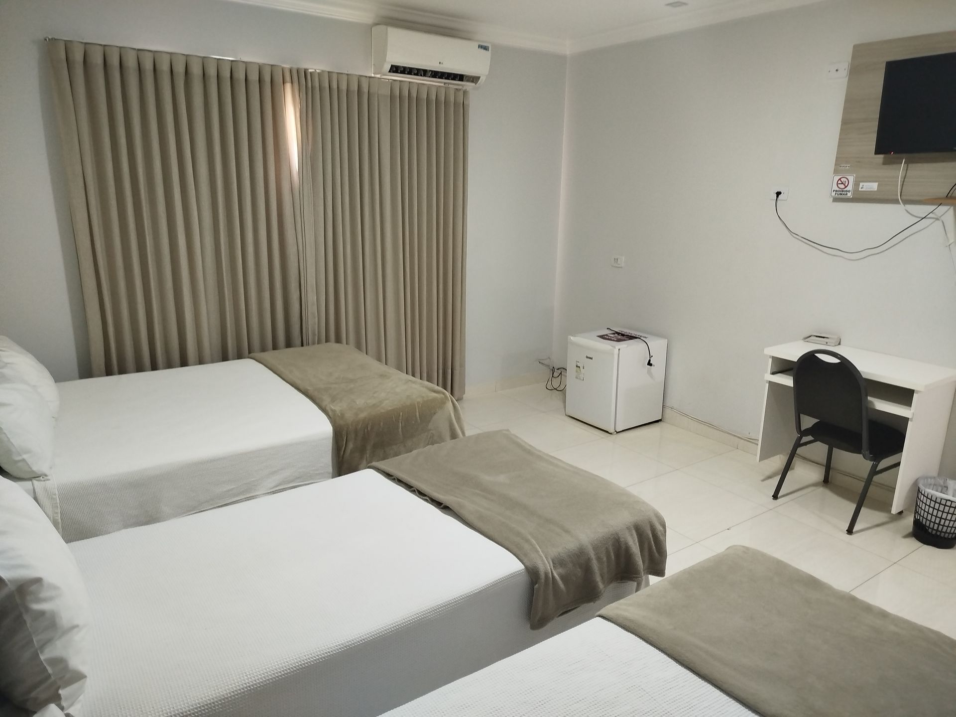 Quarto de hotel com três camas, uma escrivaninha, uma minigeladeira e cortinas.