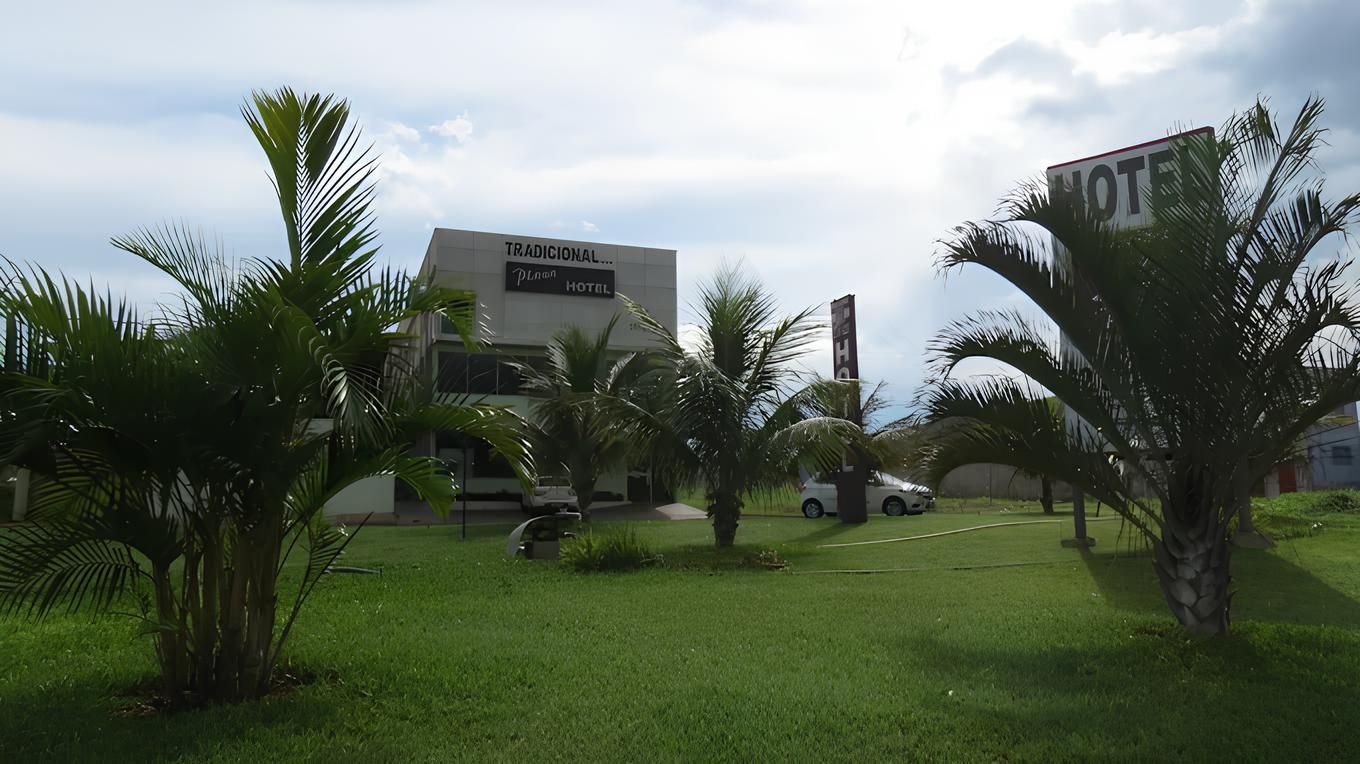 Motel com palmeiras na frente; carro branco estacionado; céu nublado.