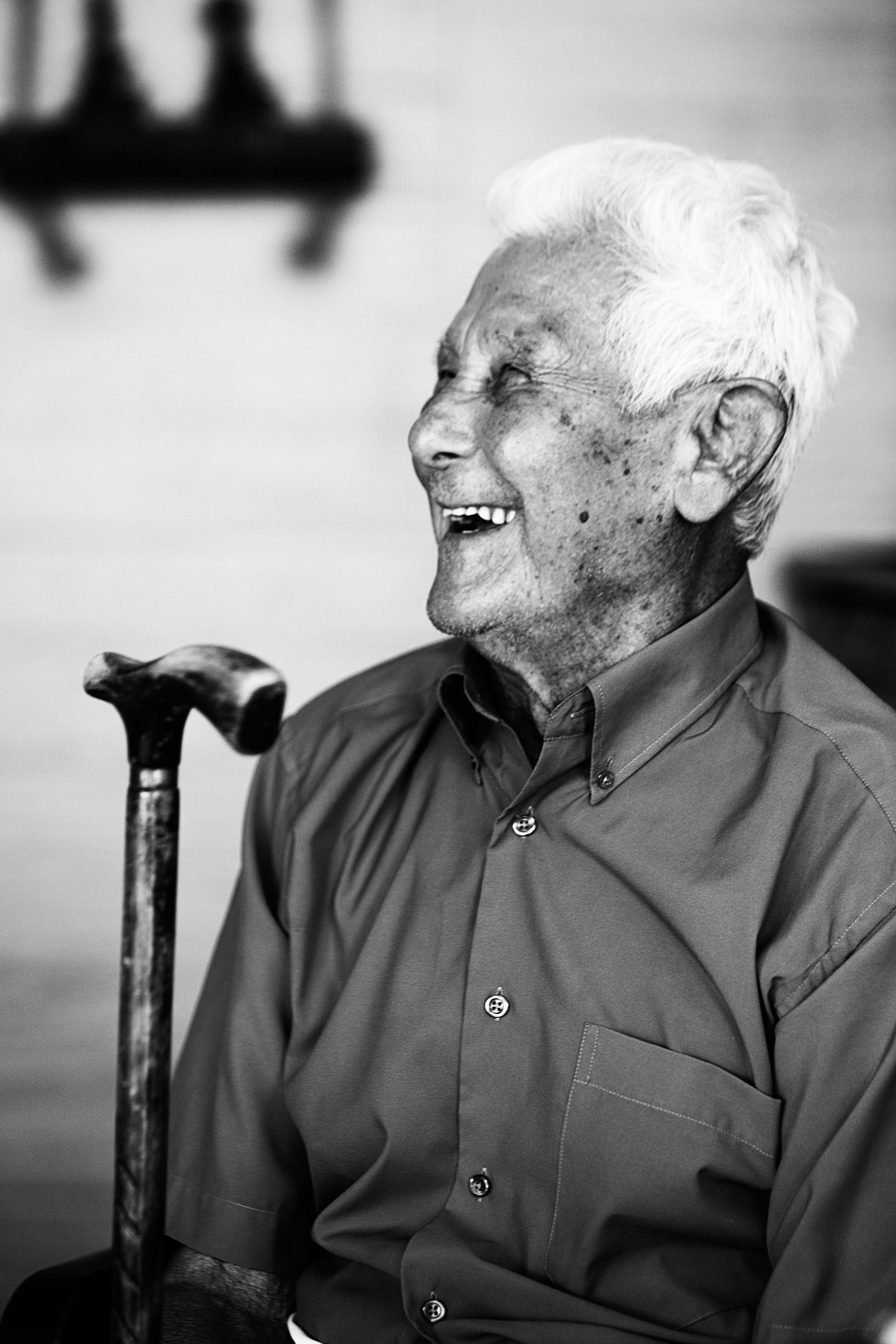 A laughing person wearing a collared shirt, sitting with their hand resting on the handle of a cane.
