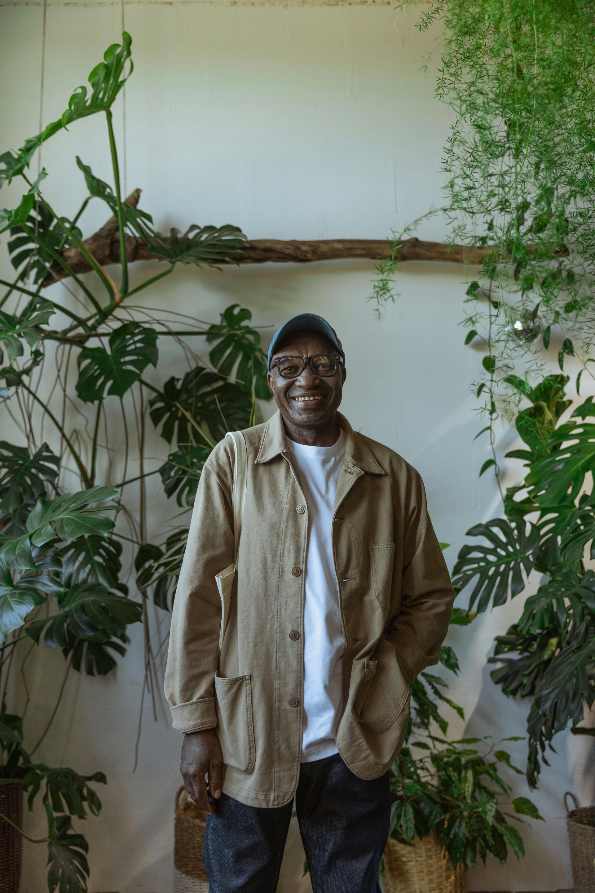 Man standing in front of lush plants, wearing a light jacket and cap, smiling.