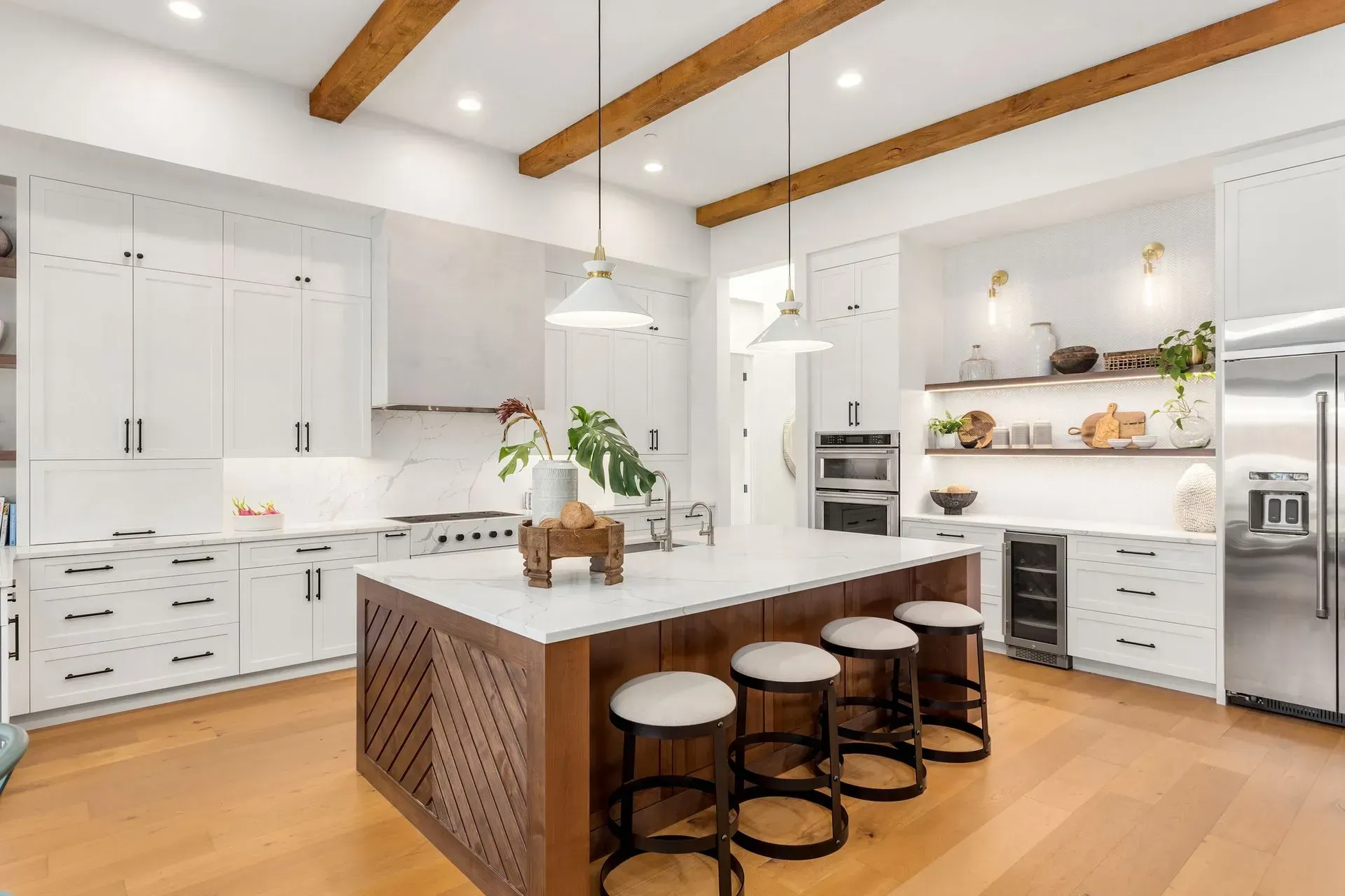 A bright, modern kitchen with white cabinets, a large wooden island with bar stools, and exposed wooden ceiling beams.
