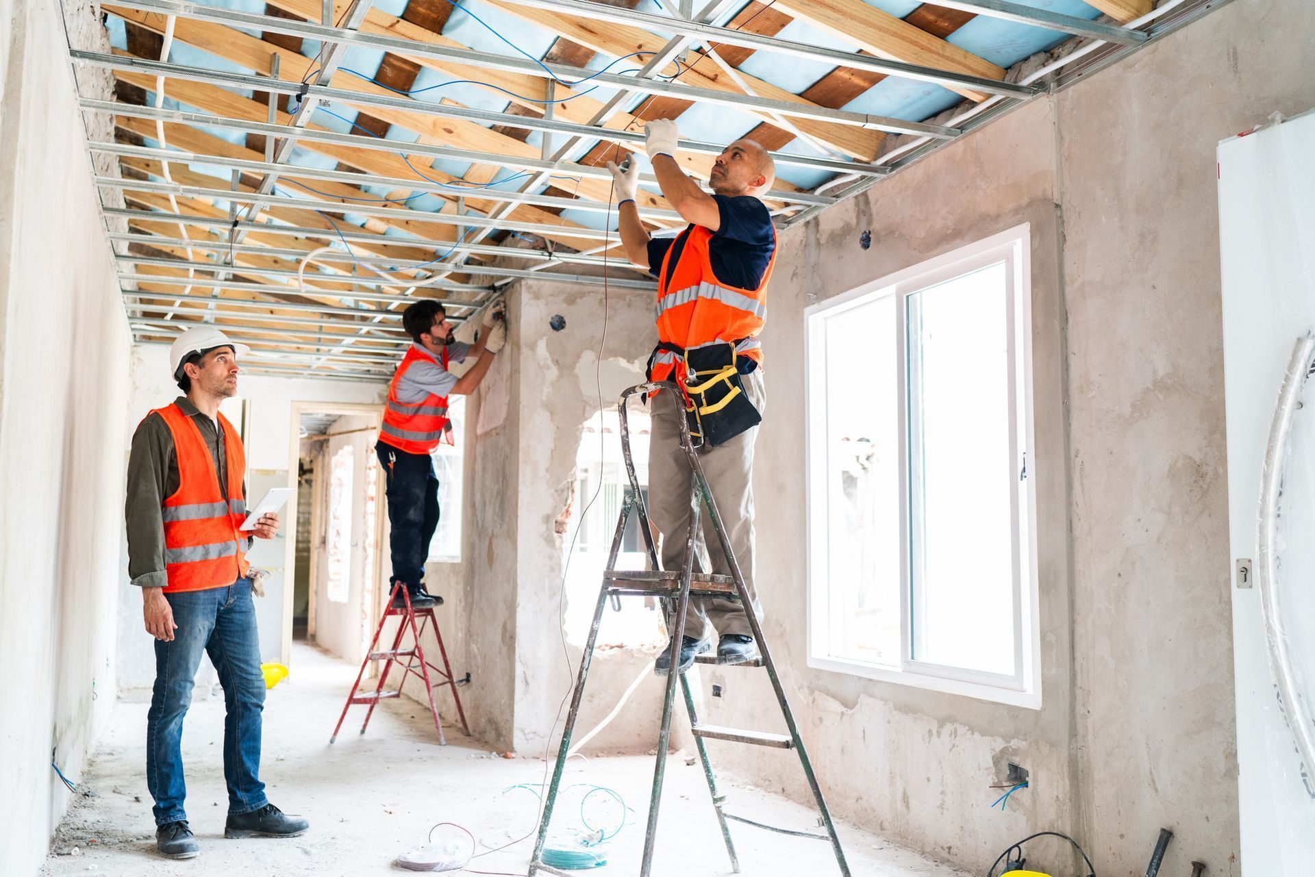 Construction workers in safety vests installing a metal ceiling frame in an unfinished room.