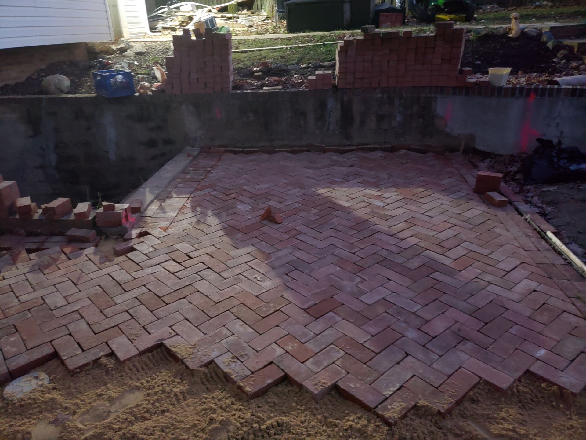 A brick patio in herringbone pattern under construction, with loose bricks, sand, and an unfinished retaining wall.