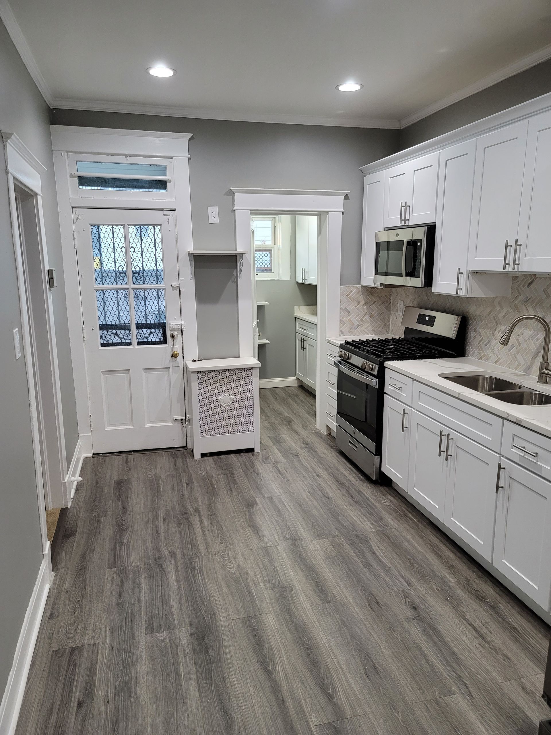 A modern galley kitchen with white cabinets, stainless steel appliances, a sink, and grey wood-look flooring.