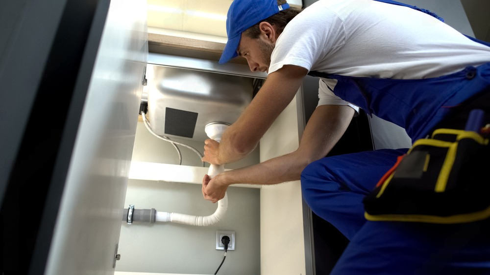 Plumber in blue uniform tightening pipes under a sink in a kitchen cabinet.