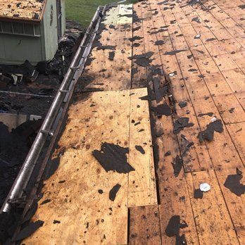 Roof with damaged asphalt shingles and exposed plywood boards, ladder visible.