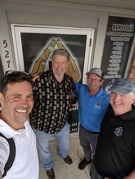 Four men smiling, standing in front of a building with a logo that looks like hands forming a house.