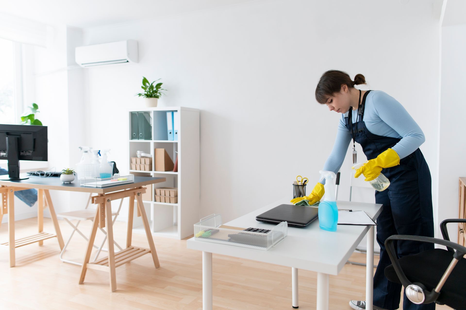 A woman is cleaning a desk in an office with a spray bottle.