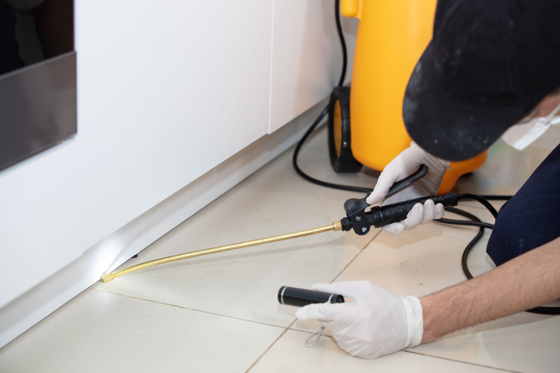 Person in gloves sprays insecticide along a floorboard near a white cabinet.