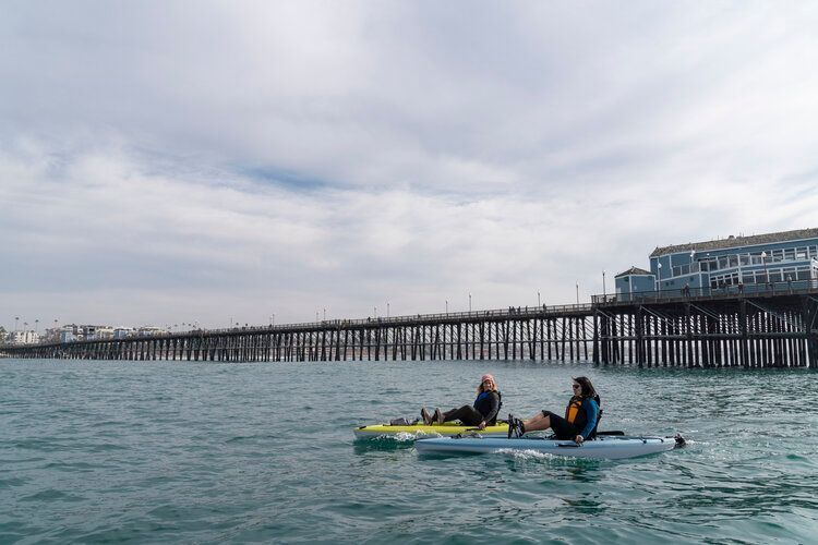 Two people are riding kayaks in the ocean in front of a pier.