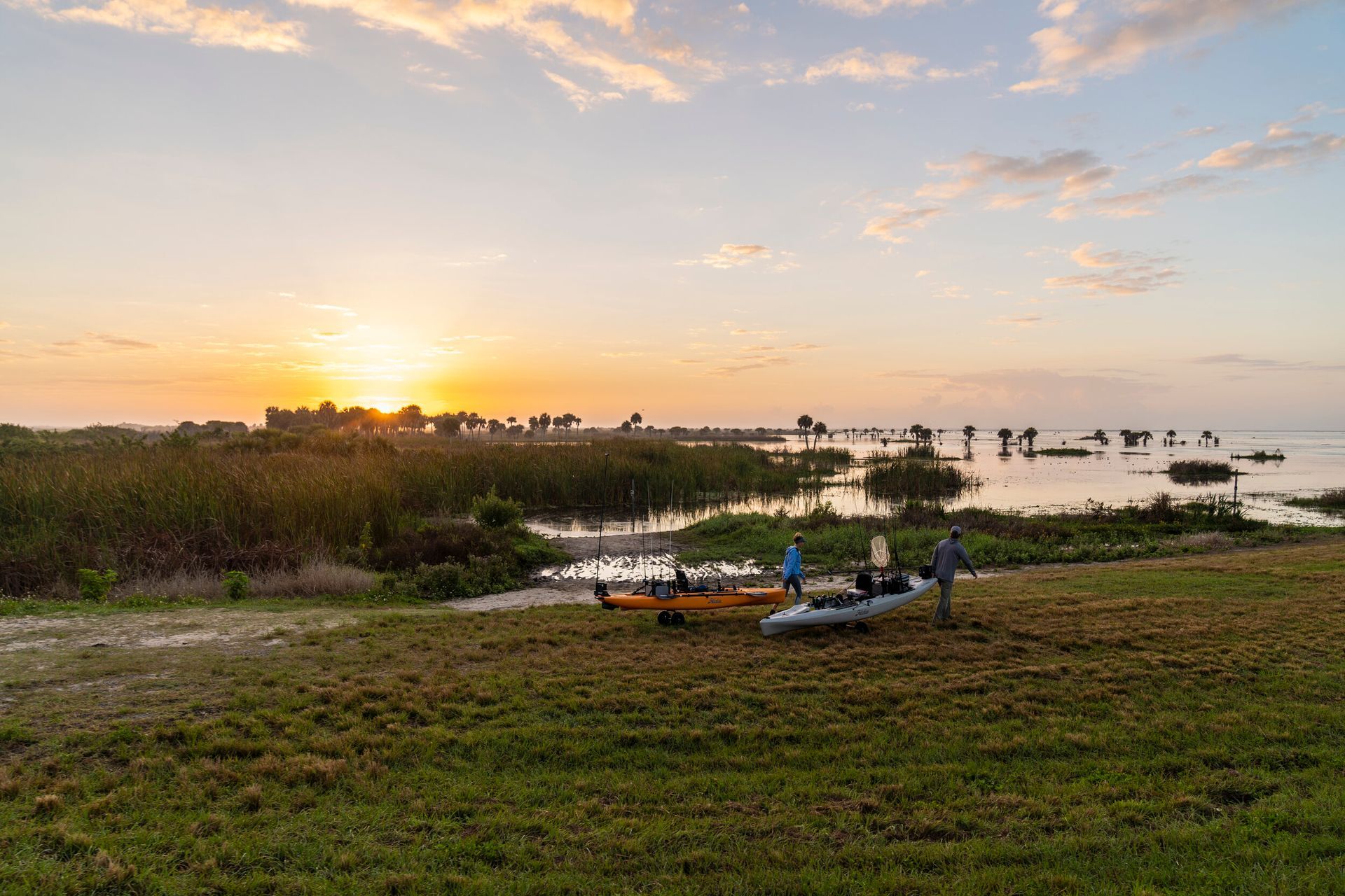 A group of people are standing in a field with kayaks in front of a body of water at sunset.