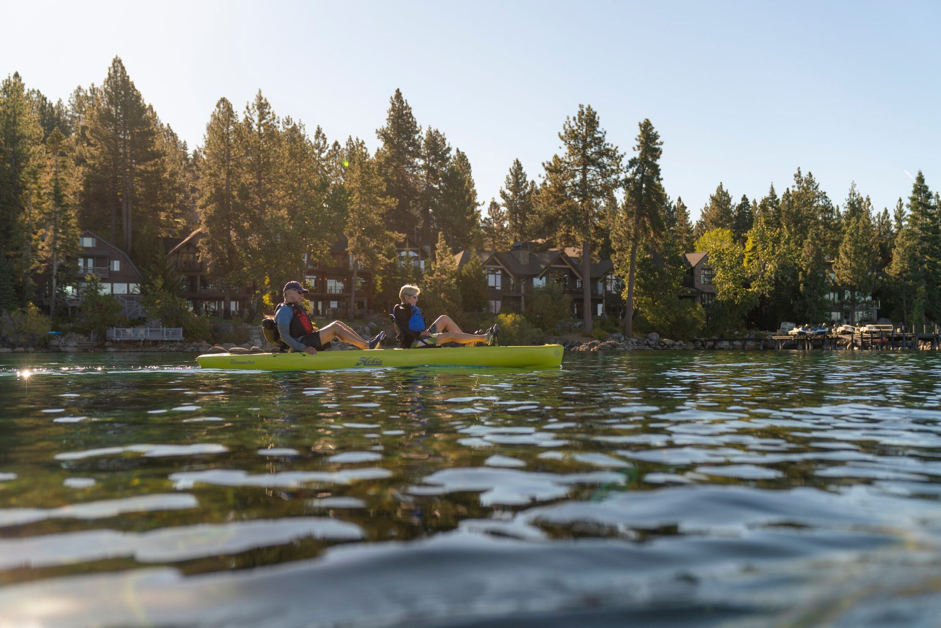 Two people are kayaking on a lake with trees in the background.