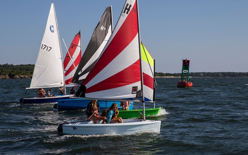 A group of people are sailing boats on a lake and one of the boats has the letter h on the sail