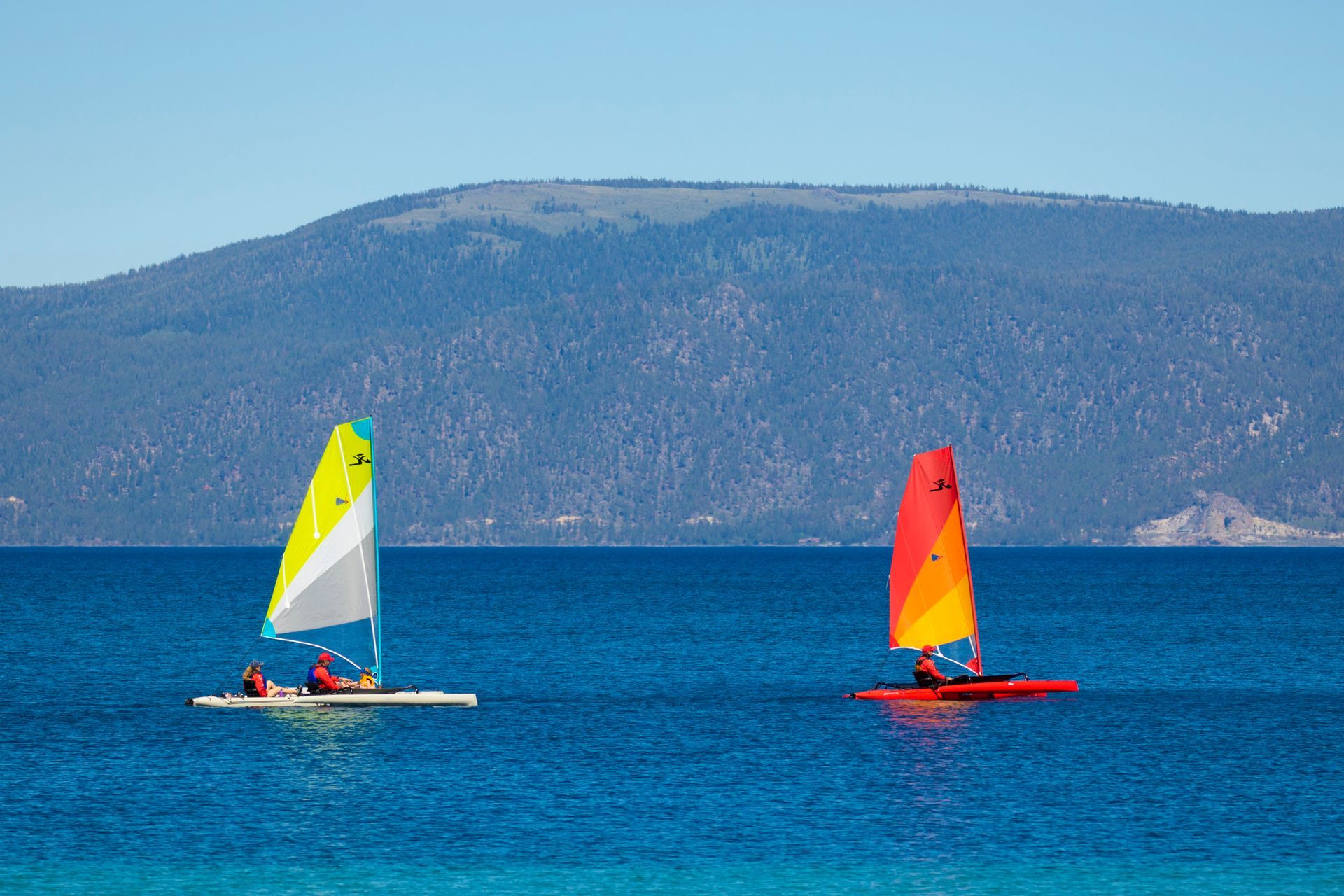 Two people are sailing boats in the ocean with mountains in the background.