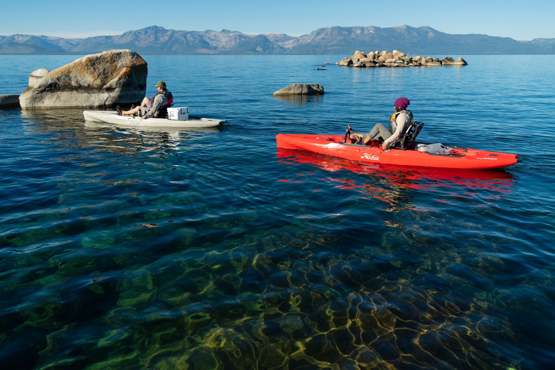 Two people are kayaking on a lake with mountains in the background.