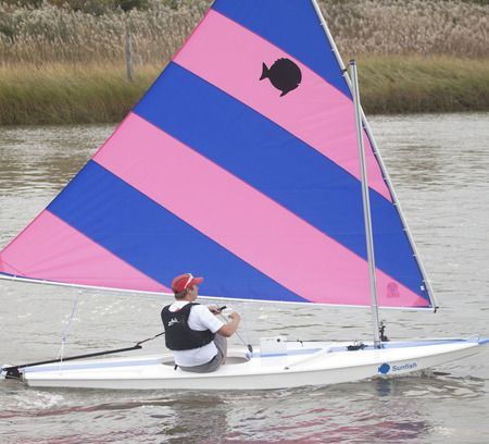 A man sits on a small sailboat with a pink and blue sail