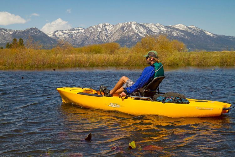 A man is sitting in a yellow kayak on a lake with mountains in the background.