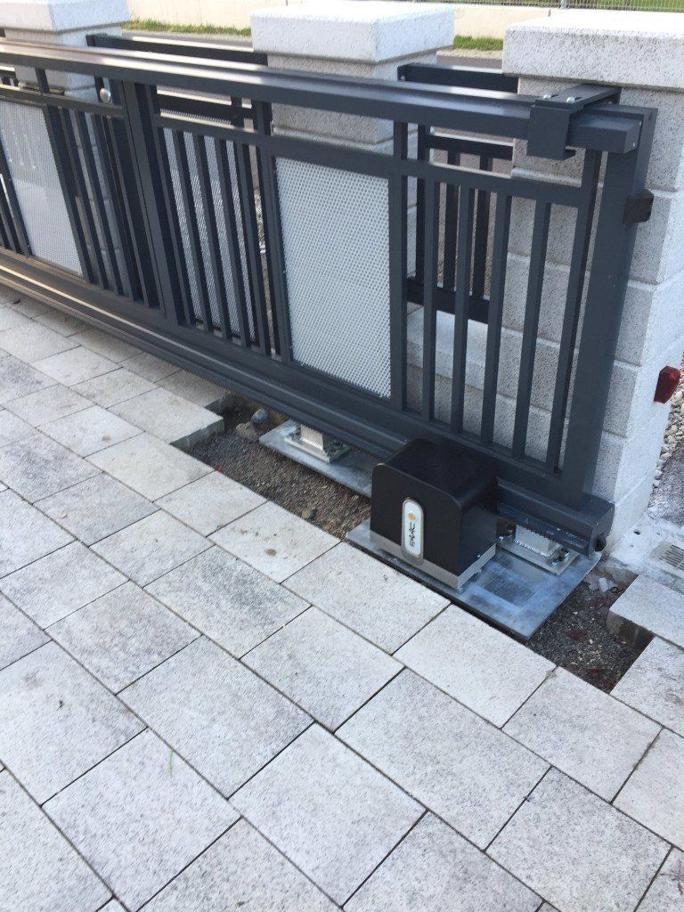 Auf einem gepflasterten Gehweg steht ein Schiebetor mit einem daran befestigten Motor.