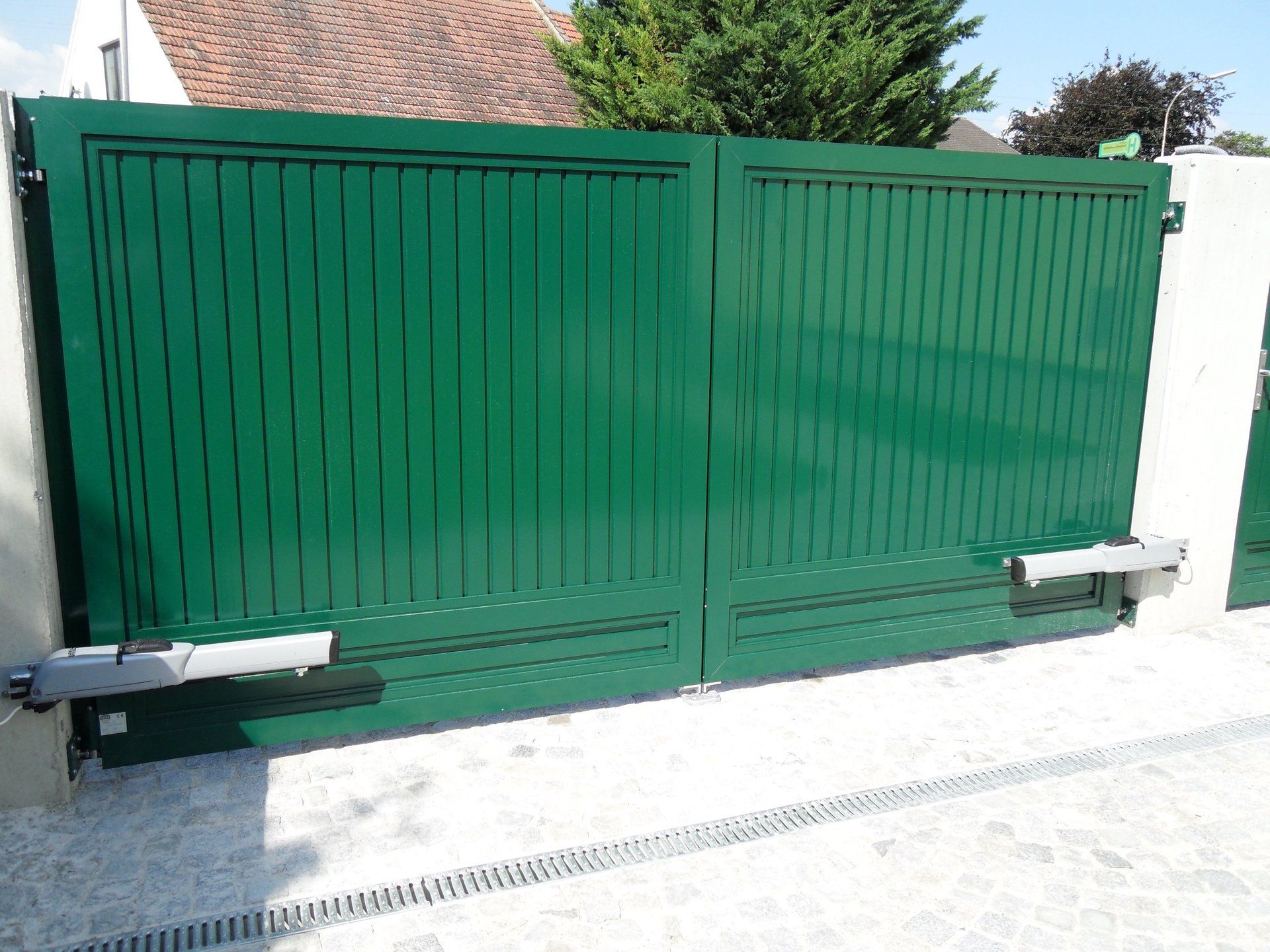 Ein grünes Tor öffnet sich zu einer Einfahrt, im Hintergrund ist ein Haus zu sehen.