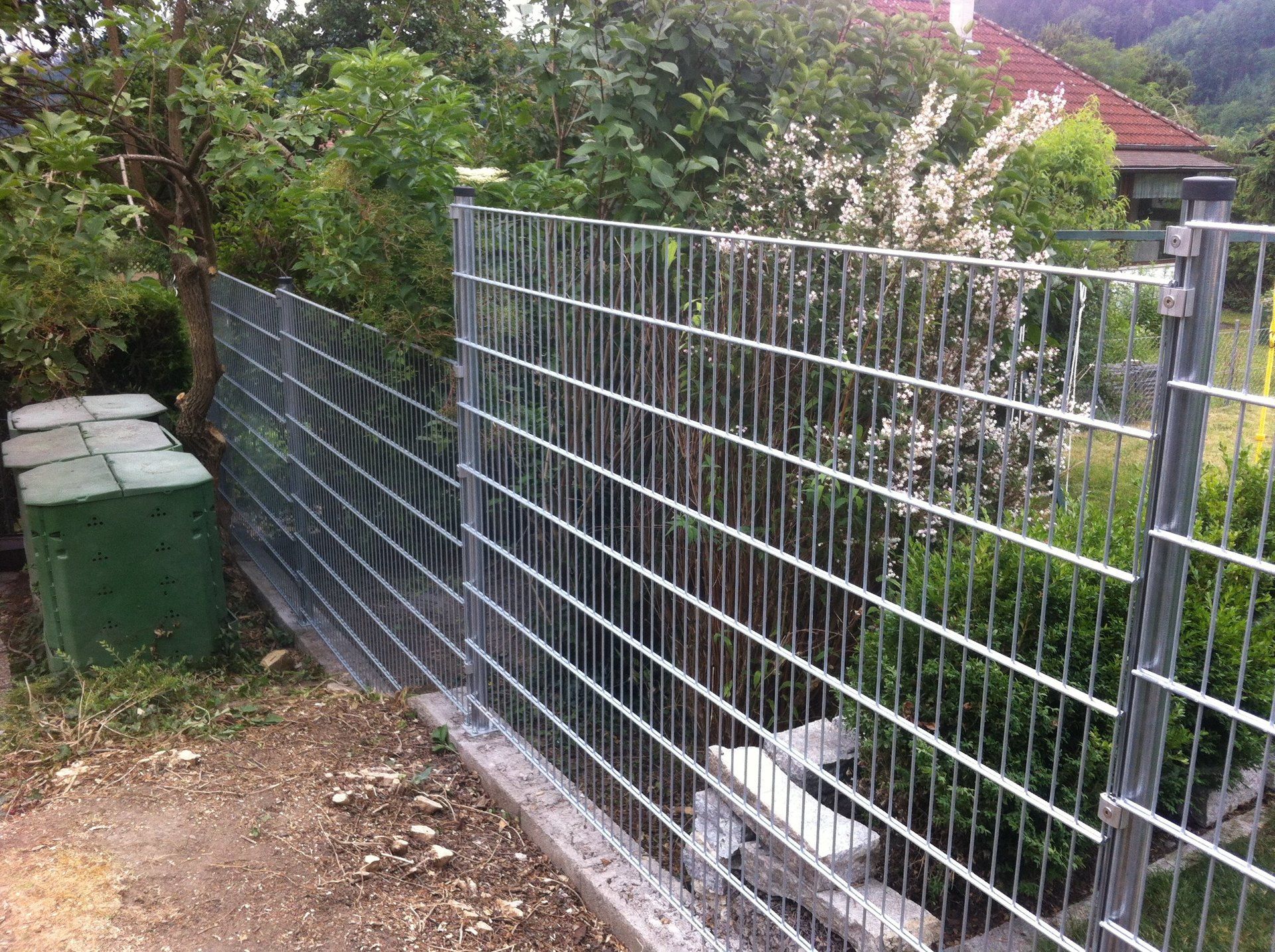 Ein Metallzaun mit einer grünen Mülltonne im Hintergrund.