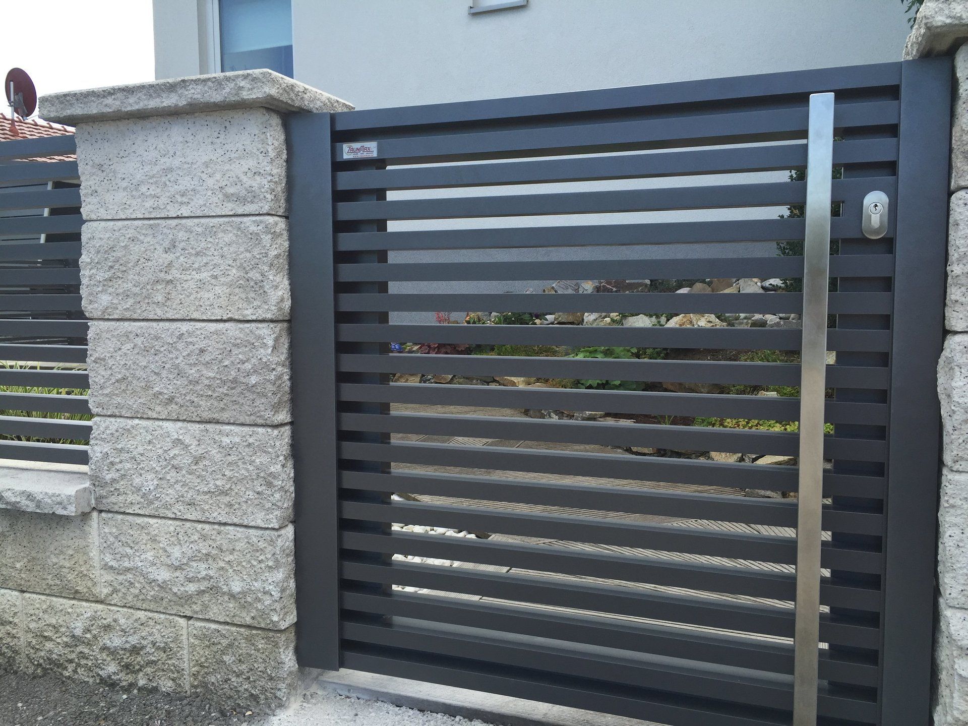 Neben einer Backsteinmauer steht ein schwarzes Tor mit einem Edelstahlgriff.