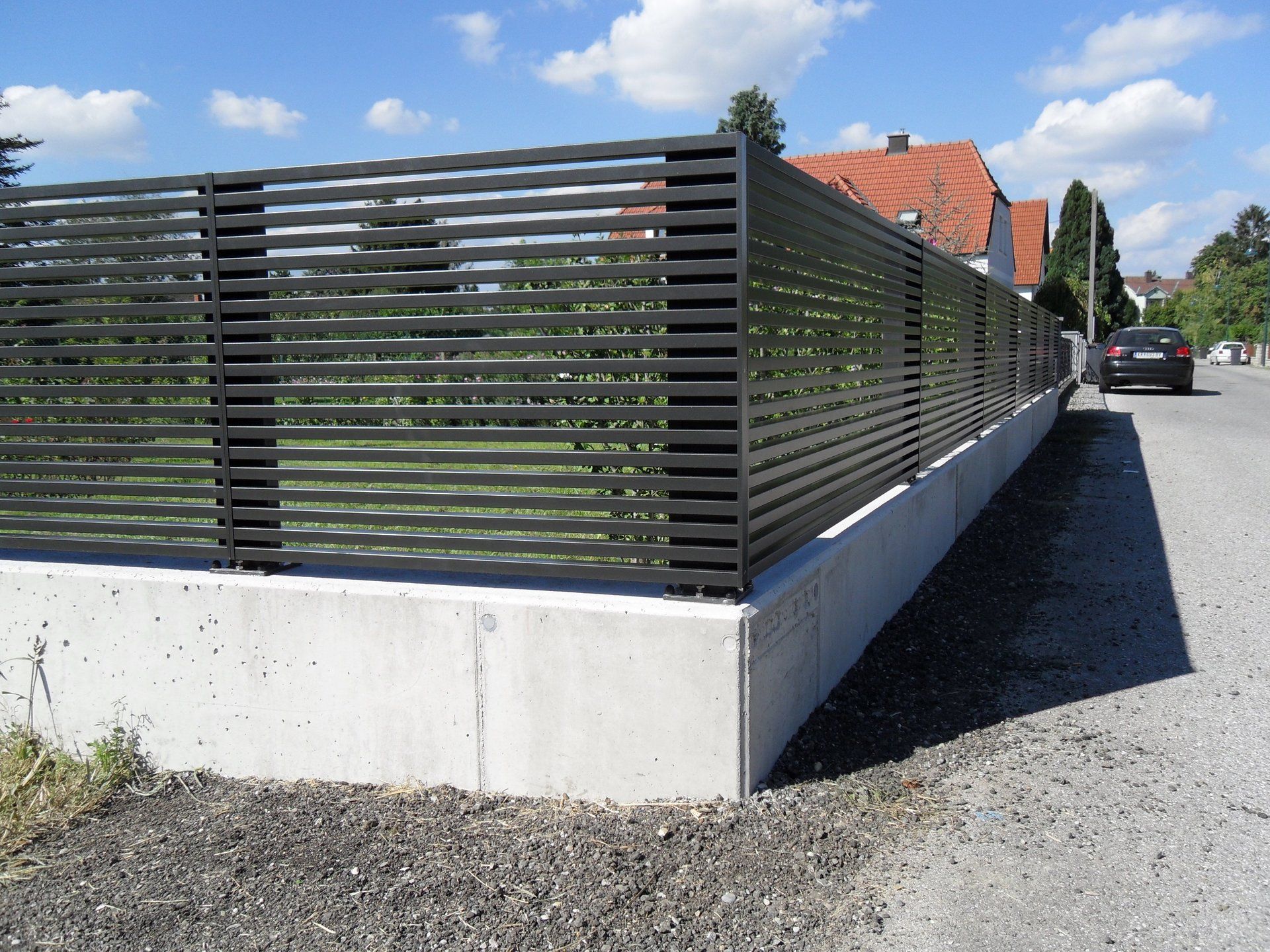Ein schwarzer Zaun entlang einer Schotterstraße mit einem dahinter geparkten Auto