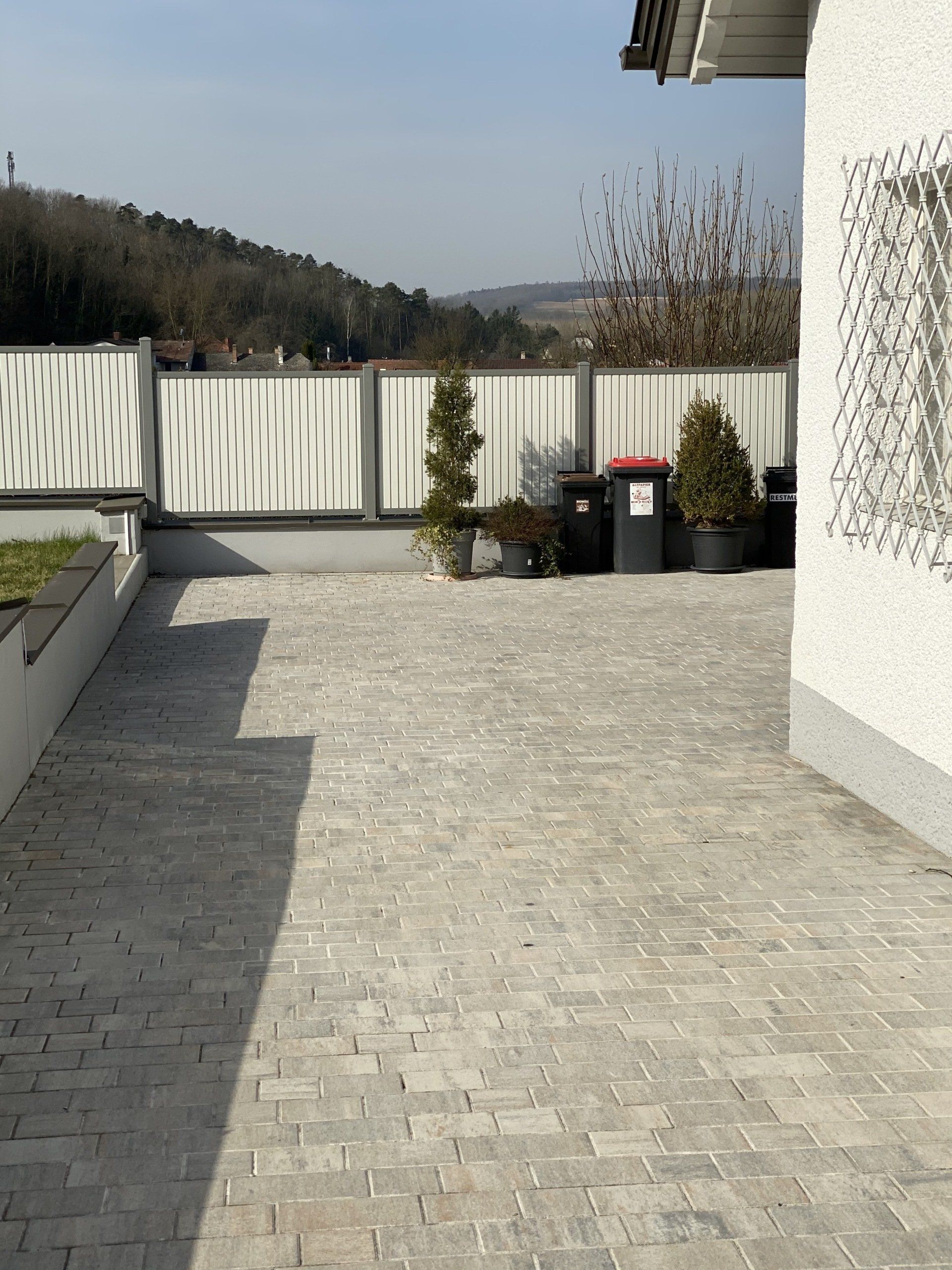 Eine gemauerte Auffahrt mit einem weißen Zaun und Mülltonnen