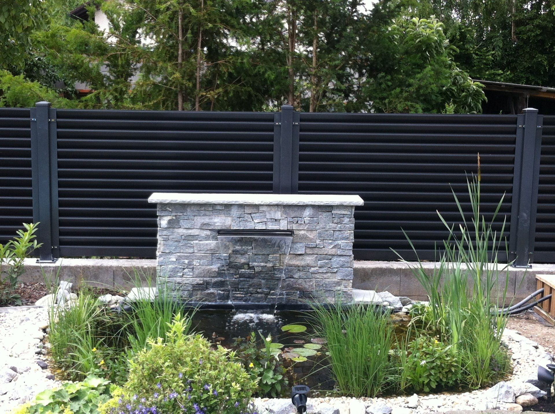 Ein Garten mit einem Teich und einem Brunnen vor einem schwarzen Zaun.