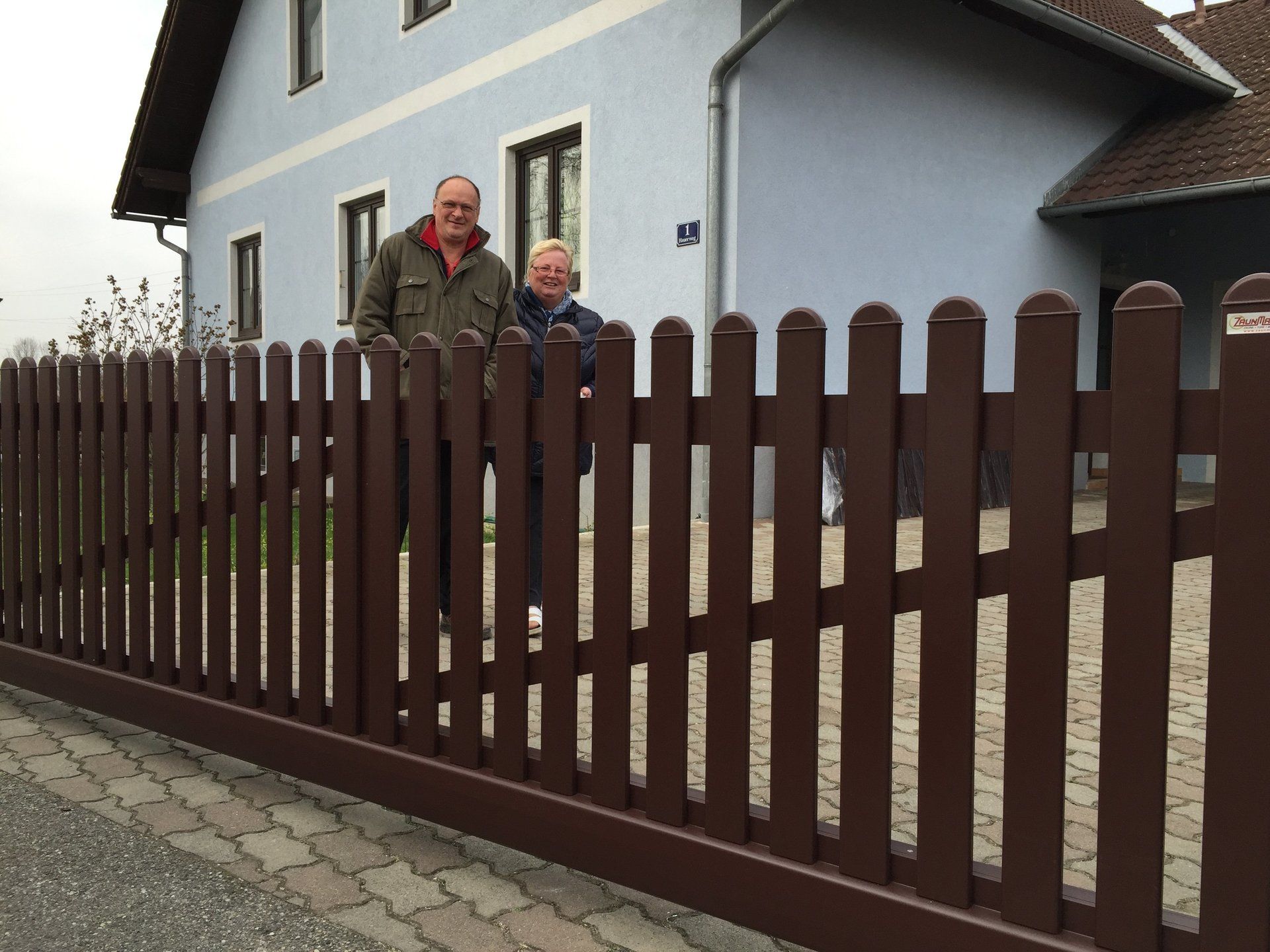 Zwei Personen stehen hinter einem braunen Zaun vor einem Haus