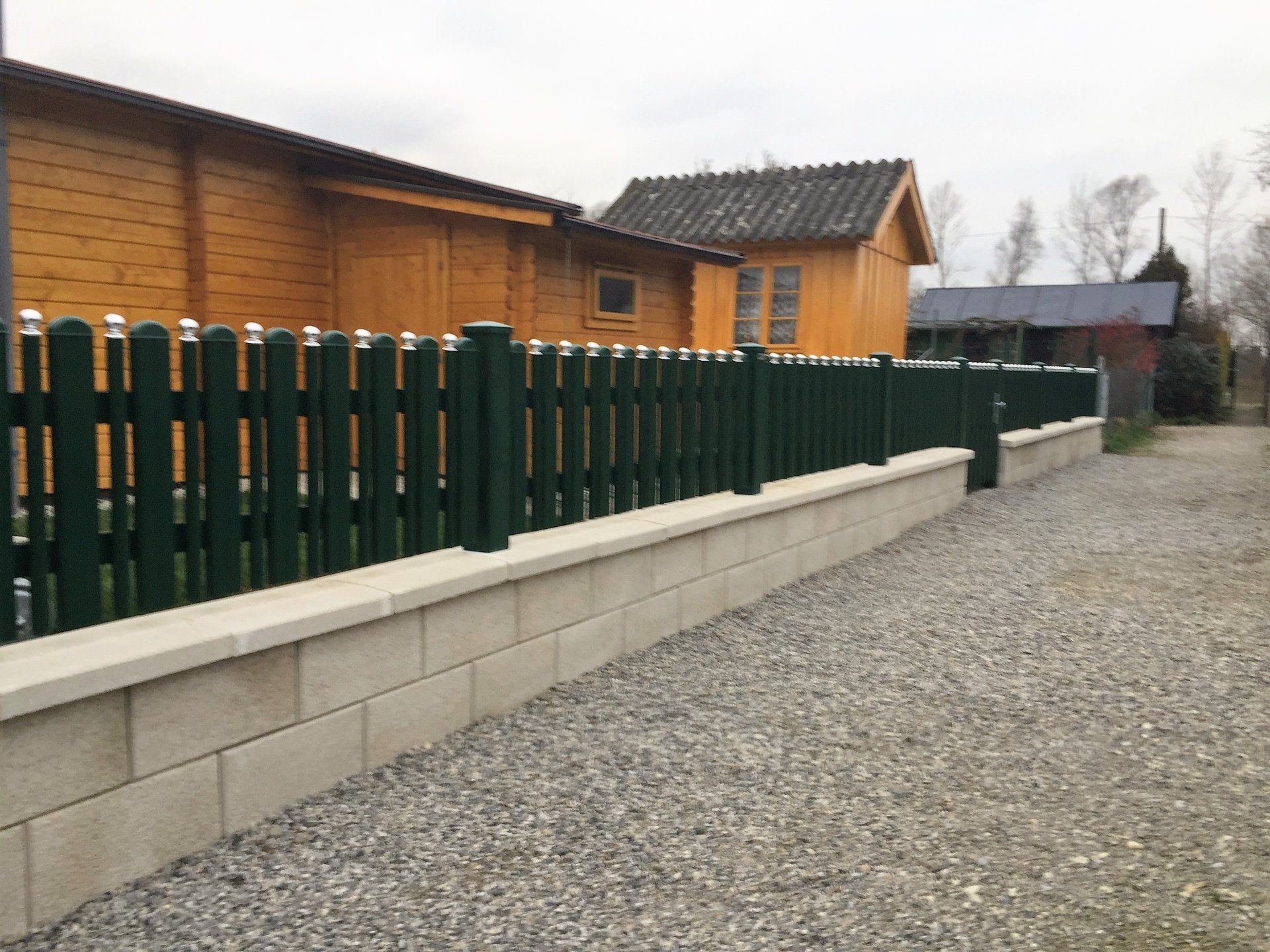 Ein grüner Zaun umgibt eine Kiesauffahrt vor einem Holzhaus.