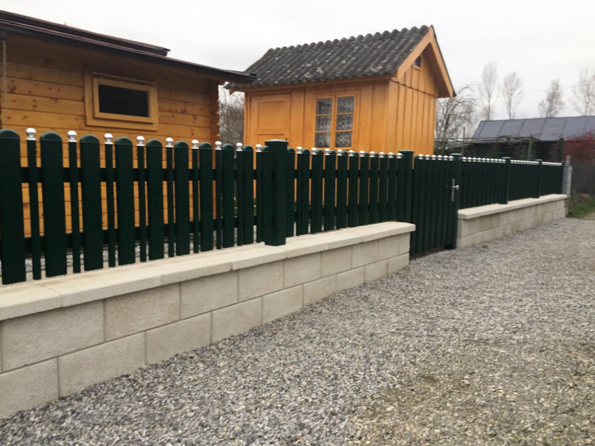 Hinter einem Zaun mit Tor steht ein Holzhaus.