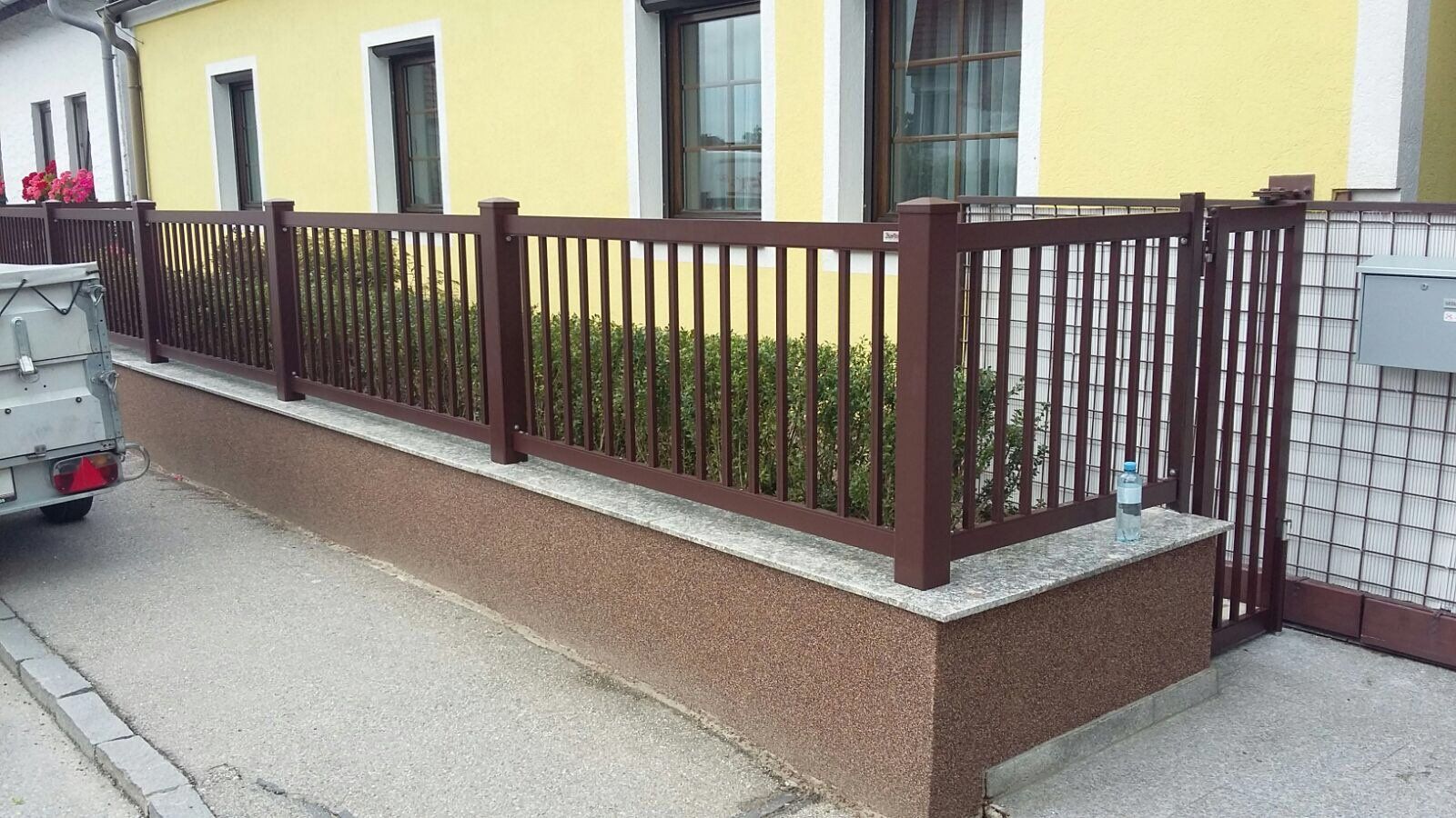 Vor einem gelben Haus steht ein brauner Zaun.