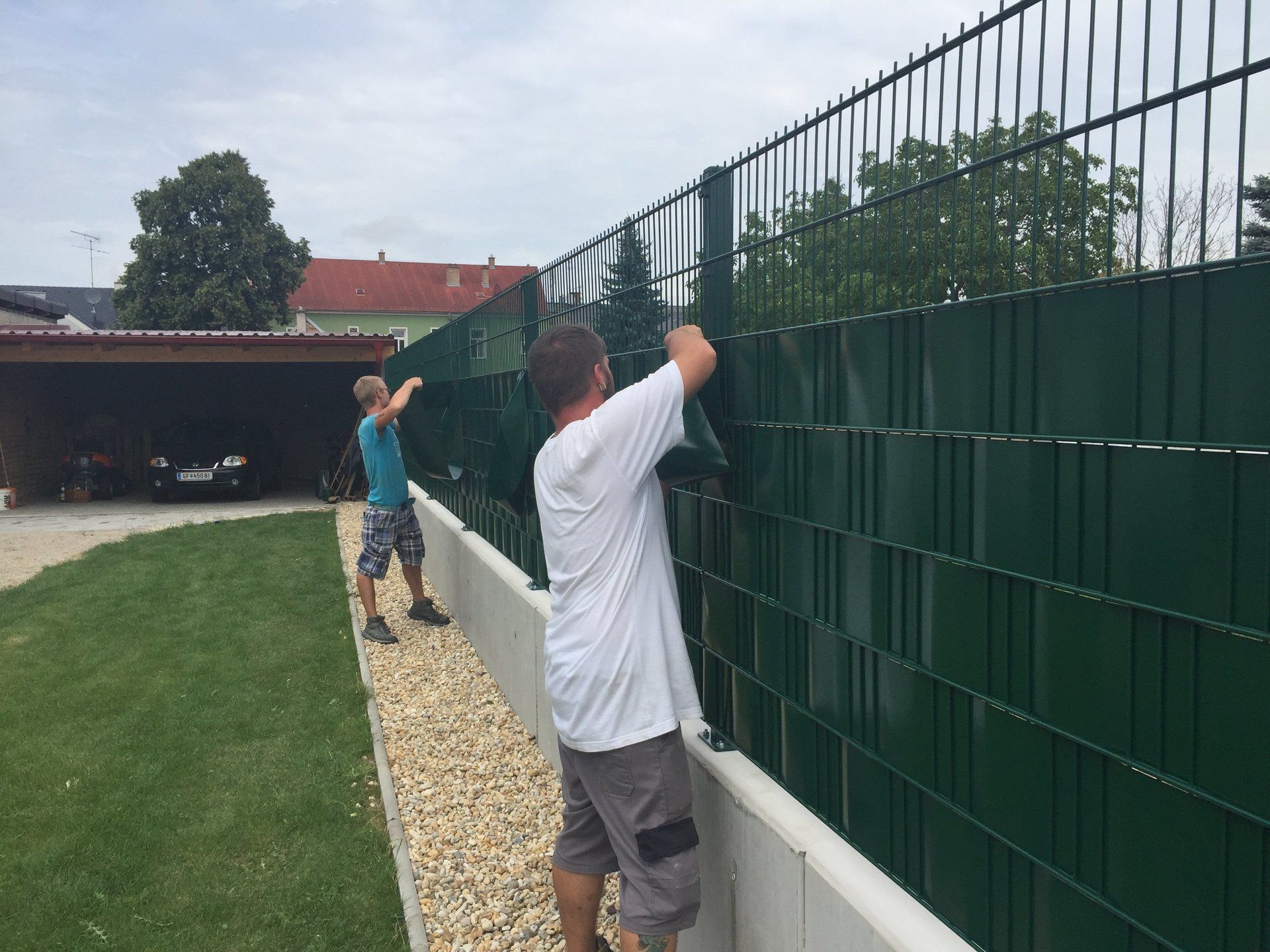 Zwei Männer installieren in einem Hinterhof einen grünen Zaun.