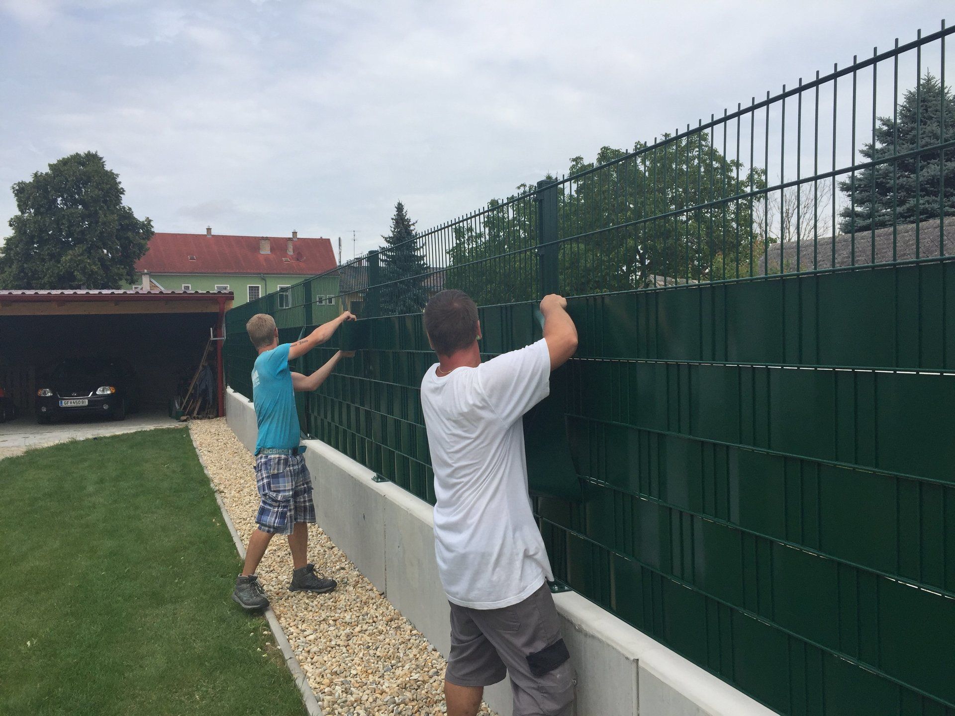 Zwei Männer arbeiten in einem Hinterhof an einem grünen Zaun.
